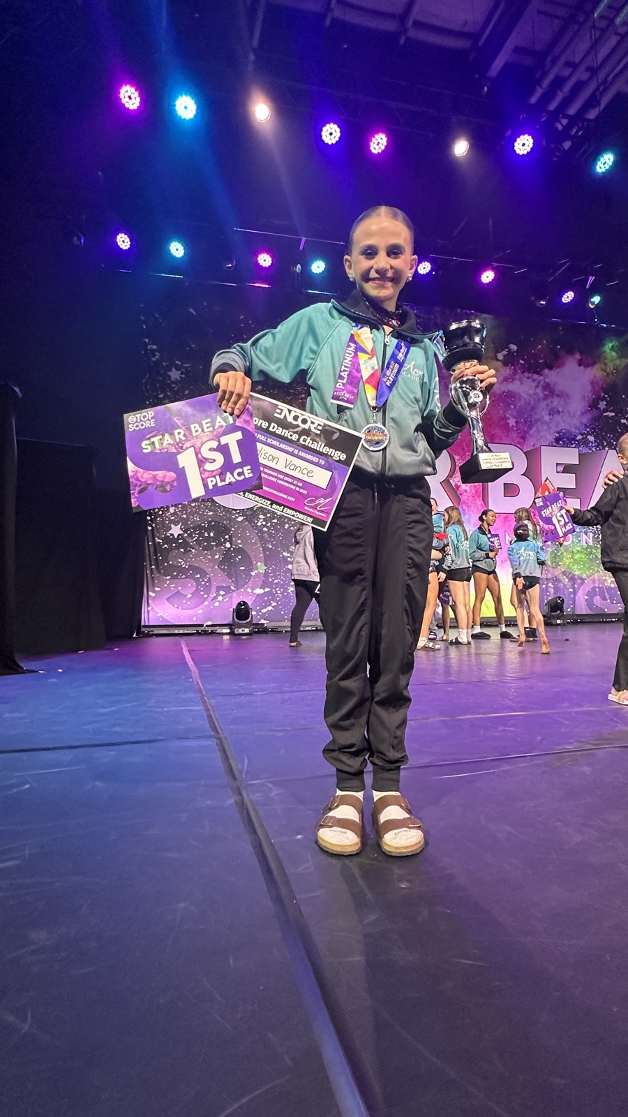 Madison Vance holding a 1st place sign, Platinum medal, and trophy on stage